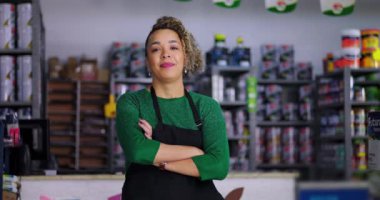 Tezgahta duran gülümseyen hırdavatçı, kollar çapraz, boya kutularıyla dolu raflar ve çeşitli renkli malzemelerle çevrili, hoş karşılama tavrı