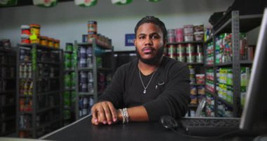 Boyacı tezgahındaki adam elleri kelepçeli, siyah önlüklü, renkli ürün raflarıyla çevrili, güven ve profesyonellik saçıyor.