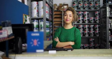 Kadın girişimci, bir hırdavat dükkanının tezgahında güvenle duruyor, kolları çapraz gülümsüyor, profesyonel bir ortamda boya ve malzeme raflarıyla çevrili.