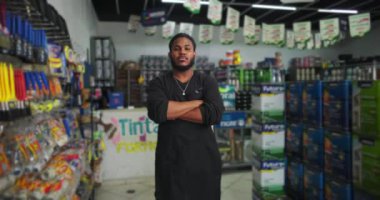 Siyah önlüklü bir adamın tüm vücudu boya kutuları ve renkli malzemelerle çevrili bir boya mağazası koridorunda kendinden emin bir şekilde dikilirken etrafa profesyonellik ve sakinlik saçıyor.