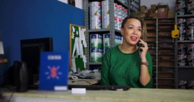 Genç bir kadın, hırdavatçıdaki bir masada telefonuna cevap verirken gülümsüyor. Etrafı ürünlerle çevrili profesyonel iletişimle meşgul oluyor.