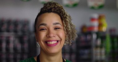 Gülümseyen dükkan sahibi bir hırdavatçıda kameraya bakıyor, arka planda renkli ürünlerle dolu raflar, pozitif bir iş ortamını resmediyor.