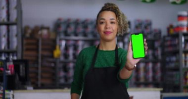 Dükkan sahibinin elinde yeşil ekran ve krom anahtar olan bir akıllı telefon var. Cihazı kameraya doğru gösteriyor. Arka planda renkli ürünler rafları var.