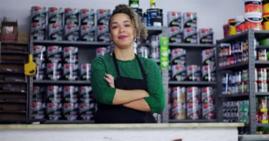 Hırdavatçıda tezgahın arkasında duran kendine güvenen kıvırcık saçlı, kollarını kavuşturmuş, gururla gülümseyen, arka planda raflarda duran boya kutuları olan genç bir kadın.