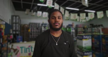 Arka planda bulanık boya ve malzeme raflarıyla çevrili bir boya dükkanında duran siyah önlüklü bir adamın yakın çekimi.