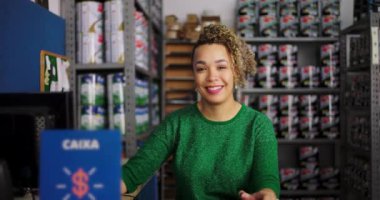 Hırdavatçıda masa başında oturan kadın girişimci, çalışırken güvenle gülümsüyor, profesyonel bir ortamda etrafını boya ve malzemelerle donatıyor.