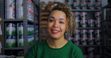 Perakende mağazasında gülümseyen bir kadın, boya kutularıyla dolu rafların önünde duruyor, iş ortamında pozitifliğini ve profesyonelliğini ifade ediyor.