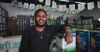 Gülümseyen tezgahtar bir hırdavatçıda istiflenmiş boya kutularına yaslanıyor, canlı, ürün dolu bir çalışma alanında müşteri dostu hizmet sunuyor.