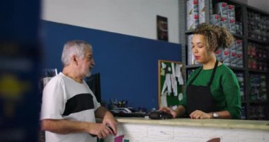 Gülümseyen genç bir kadın ve yaşlı bir adam hırdavatçı dükkanındaki alışveriş sırasında neşeli bir takasa giriyorlar.