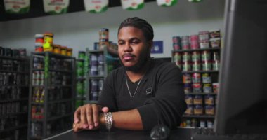 Boya dükkanındaki kasaya dayanan düşünceli bir esnaf. Etrafı boya kutuları ve iş yerindeki malzemelerle çevrili.