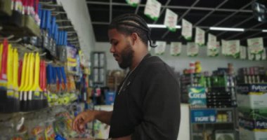 Siyah önlüklü bir adam boya dükkanında fırça ve aletleri sınıflandırıyor, bir fırça ve karton kutu tutuyor, müşteriler için vitrinlerde canlı malzemeler düzenliyor.