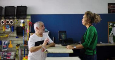 Dükkan sahibi, hırdavatçıda yaşlı bir müşteriyi dikkatle dinliyor. Müşteri ilişkilerinde sabır ve anlayış gösteriyor.