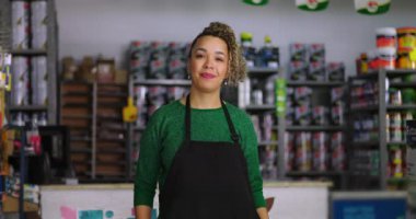 Dost canlısı genç bir kadın, bir nalburun arkasında, yüzünde bir gülümsemeyle, boya ve hırdavat malzemeleriyle dolu raflarla güvenle dikiliyor.