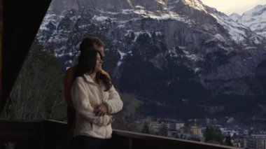 Dağ evinin balkonundan dağların manzarasını seyreden mutlu bir çift. Kadının biri ufku işaret ederken, kocası kollarını ona dolamış.