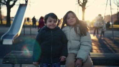 5 yaşında bir oğlan ve 8 yaşında bir kız. Park, Sunset Backlit Sahnesi 'nde. Arka planda oynayan bulanık çocuklar. Kardeşler bankta oturur ve kameraya bakarlar.