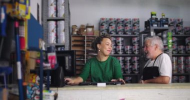 Erkek ve kadın, etrafları boya ve aletlerle çevrili küçük işletme hırdavatçısının tezgahında gelişigüzel sohbet ediyorlar. Meslektaşlar arasında dostça bir iş ilişkisi var.