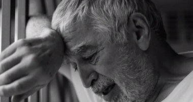 Pencere parmaklıklarına yaslanmış yaşlı bir adam, ezici üzüntüyle boğulmuş, gözleri sessiz bir umutsuzluk içinde kapalı, kapalı bir yerde derin bir duygusal acıyı yakalıyor, siyah ve beyaz.