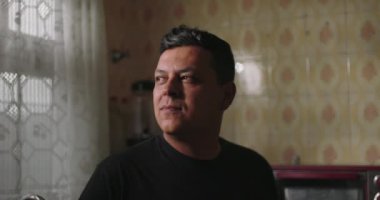 Mutfak penceresinin önünde düşünceli bir şekilde duran adam, etrafı dantelli perdeler, fayanslı duvarlar ve ev aletleriyle çevrili, rahat ve klasik bir iç mekan ile çevrili.
