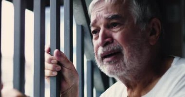 Pencere parmaklıklarını tutan yaşlı bir adam. Yüzü ıstırapla kıvrılmış. Kapalı alandaki duygusal kırılganlık anında ezici keder ve umutsuzluğu ifade ediyor.