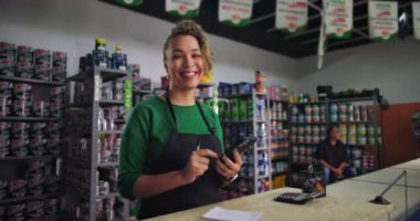 Kameraya gülümseyen kıvırcık saçlı küçük bir işletme sahibi hırdavatçı tezgahında telefon tutarken etrafı boya kutuları ve aletlerle dolu.