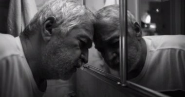 Yaşlı bir adam, alnını banyo aynasına bastırıyor. Üzüntü ve duygularının ağırlığı altında eziliyor. Hem de derinden ham ve içsel bir anda.