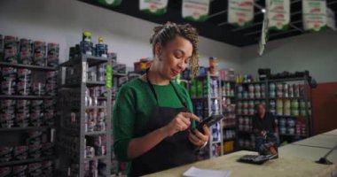 Hırdavatçıda duran kıvırcık saçlı küçük işletme sahibi, telefonuna odaklanmış bir ifadeyle bakıyor, etrafı boya kutuları ve malzemelerle dolu raflarla çevrili.