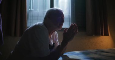 Elleri dua ederken yatakta oturan yaşlı bir adam ilahi rehberlik arıyor ve inancın yansıtıcı anını umut ve yalnızlığın içsel gözlemlerini destekliyor.