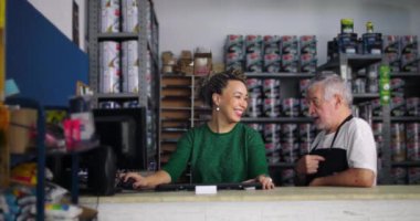 Gülümseyen adam ve kadın, yerel bir nalbur dükkanının tezgahında neşeli bir anı paylaşıyor. Etrafı boya ve aletlerle çevrili, dost canlısı bir iş ortamı yaratıyor.