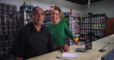 Tezgahta gülümseyen bir bayan müşteriyle birlikte duran küçük işletme sahibi. Arka planda boya kutuları ve malzemeler var. Dostça ve hoş karşılayıcı.