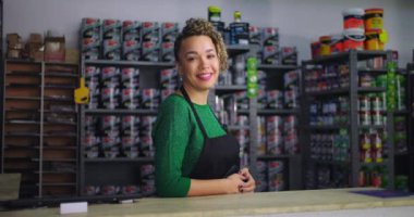 Tezgahta duran gülümseyen kadın küçük işletme nalburunda, etrafı boya ve aletlerle çevrili, işyerinde güven ve profesyonellik yayıyor.