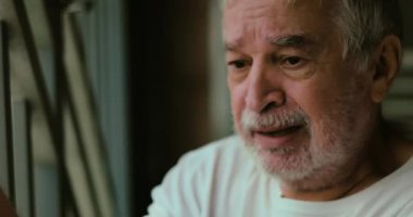 Endişeli bir ifadeye sahip yaşlı bir adam, metal parmaklıklara tutunarak çevreyi gözlüyor, yalnızlığı, ve ihtiyarlığın iç yüzünü temsil ediyor.