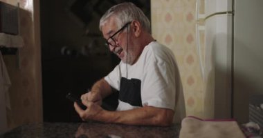 Yaşlı adam mutfak masasında akıllı telefon kullanırken gülümsüyor, teknoloji ile meşgul ve rahat, sıcacık ve retro mutfak iç tasarımı ile çevrili.