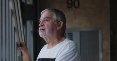 Metal parmaklıklara yaslanmış yaşlı bir adam, izole edilmiş bir garajdan sokağı izliyor ve izliyor, bir anlık bir yansıma, yalnızlık ve düşünceli olma anı yakalıyor.