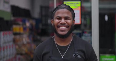 Önlük giymiş gülümseyen genç siyahi adam, perakende mağazasının önünde güvenle duruyor, bir işadamı ya da işçi olarak rolünden olumlu ve gurur duyuyor.