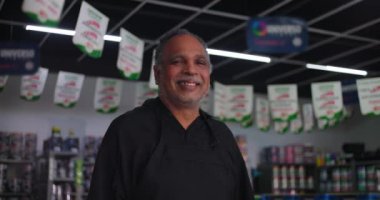 Gülümseyen küçük işletme sahibi kendine güveni tam bir şekilde nalbur dükkanında, tavandan sarkan pankartlar ve arka planda boya kutuları ve erzak rafları