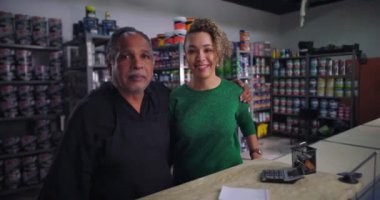 Baba ve kızı, ailelerinin hırdavat dükkanında tezgahın arkasında yan yana duruyorlar. Arka planda boya kutuları ve malzemeler var. Gururlu ve dost canlısı bir ortam.