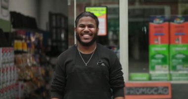 Gülümseyen genç bir siyah adam perakende mağazasının önünde duruyor, rahat duruş, pozitiflik, güven ve profesyonel ortamda yaklaşılabilir davranış.