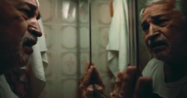 Yaşlı bir adam ellerini aynaya kaldırıyor, geçmişinin acısıyla yüzleşirken sessizce cevaplar için yalvarıyor ve duygusal bir sahnede ezici bir suçlulukla güreşiyor.