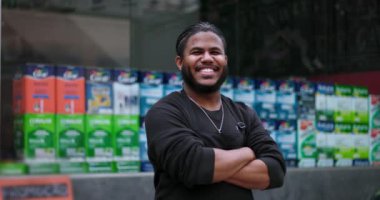 Gülümseyen genç siyahi adam kollarını kavuşturup mağazanın önünde duruyor. Profesyonel bir ortamda güven, pozitiflik ve liderlik yayıyor.