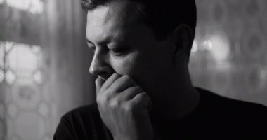 Dramatik siyah beyaz giyen, başı öne eğilmiş, elleri boyunda, derin düşüncelere dalmış, ciddi bir atmosferde endişe ve iç karışıklığı çağrıştıran bir adam.