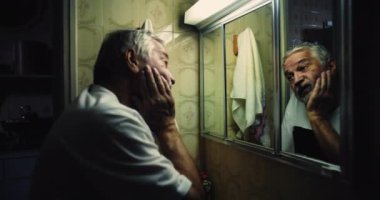 Banyodaki aynada yansımasına yaslanmış yaşlı bir adam. Yoğunlukla dolu gözler. Sanki kayıp zamanı arıyor ve kendi varlığıyla boğuşuyor.