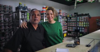 Baba ve kızı, ailelerinin hırdavat dükkanında yan yana duruyorlar. Arka planda boya kutuları ve malzemeler var. Birlik ve gurur gösteriyorlar.