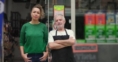 Kıvırcık saçlı kadın müşteri ve yaşlı erkek esnaf bir mağazanın önünde yan yana duran önlük giyiyorlar. Hem ciddi hem de kendinden emin ifadeleri var.