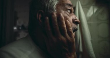 Yaşlı bir adam, kederle ve umutsuzlukla boğulmuş bir şekilde yüzünü ellerinin arasına alıyor. O, sessiz bir ızdırap ve yansıma anında yukarıya bakıyor.