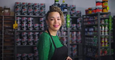 Kendine güvenen bir kadın, küçük işletme hırdavatçısında gülümsüyor, boya ve aletlerle dolu rafların önünde duruyor, profesyonellik ve misafirperver tavırlar sergiliyor.