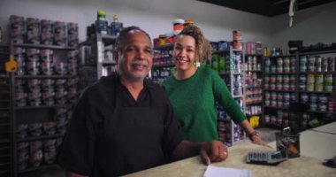 Küçük işletme sahibi tezgahta bayan bir meslektaşıyla birlikte bir hırdavatçıda gülümsüyor. Arka planda boya kutuları ve malzemeler var. Olumlu ve dostane bir atmosfer.