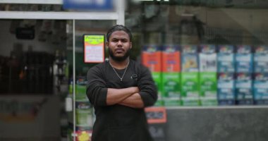 Satış mağazasının önünde kolları bağlı duran kendinden emin genç siyahi adam, iş ortamında otorite, kararlılık ve profesyonelliği yansıtıyor.