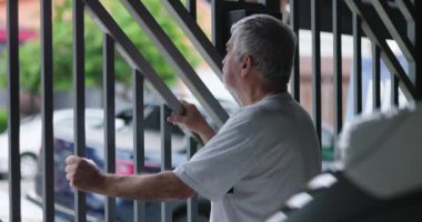 Metal parmaklıklara tutunmuş yaşlı bir adam, yansıtıcı bir ifade, izolasyon ve dış dünyayla bağlantı ile evinden sokağa bakıyor, yaşlılığın sessiz yalnızlığı.