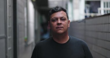 Şehir merkezinde ciddi bir adam dikiliyor, doğrudan bakıyor, yüz ifadesi sakin, şehir ortamında sakin bir an, kentsel ortamdaki kişisel yansıma