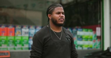 Bir odaklanmış genç siyahi adam perakende mağazasının yanında duruyor, uzaklığa bakıyor, hırsını, profesyonelliğini ve iş ortamındaki kararlılığını aktarıyor.
